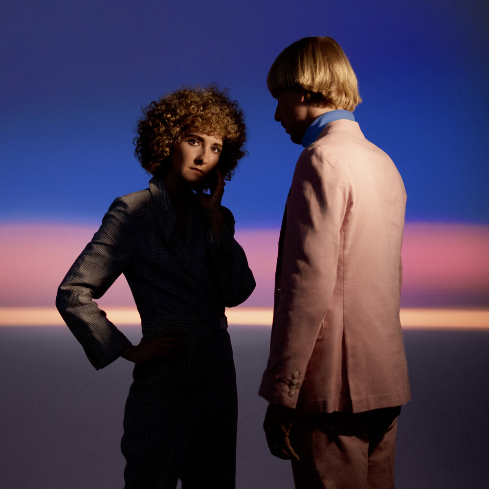 In front of a blue and purple background, Alaina Moore and her partner, Patrick Riley are standing but not looking at each other.