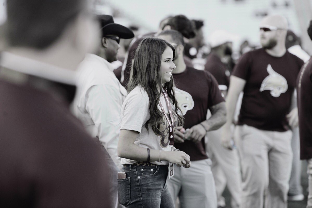 : Black and White photo of Haleigh Blocker smiling on the sidelines with other staff members in the background.
