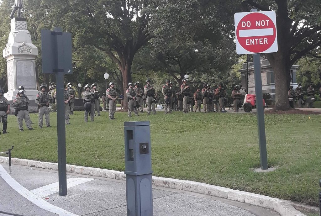 Police officers in military clothing stand lined up in downtown Austin Capitol Park in the middle of a protest.