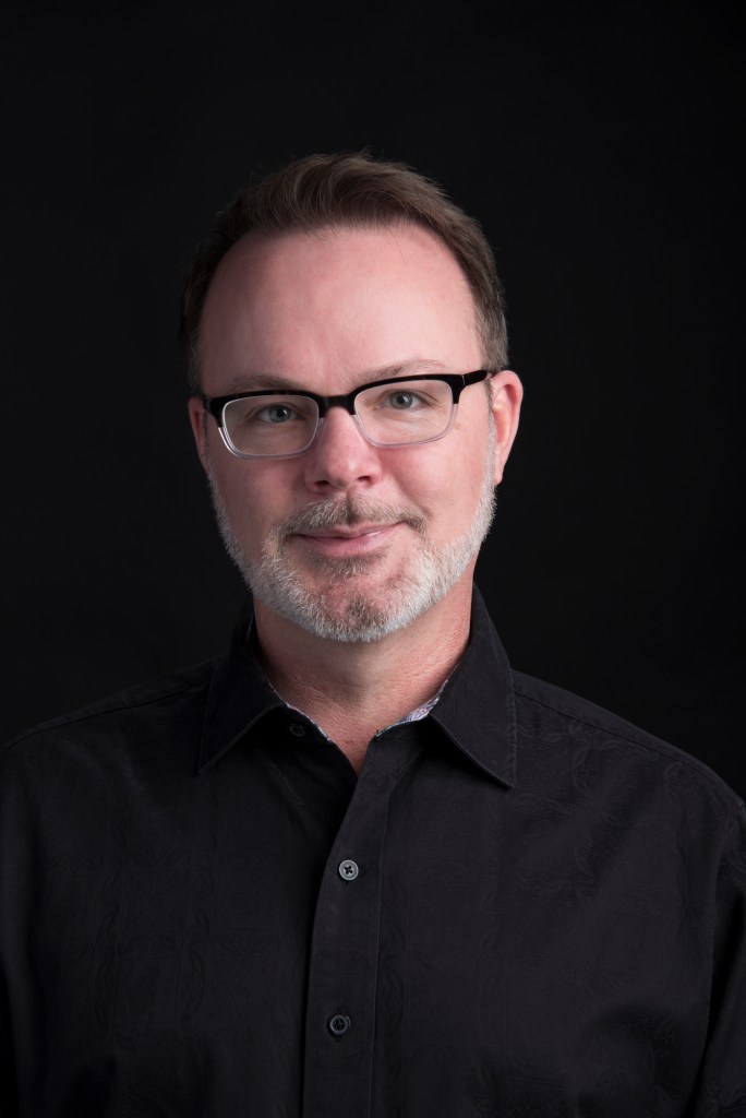 A portrait image of Texas State University's School of Music director, Dr. Joey Martin. Black background with a man wearing a solid black shirt and thick-rimmed glasses stares directly at you.