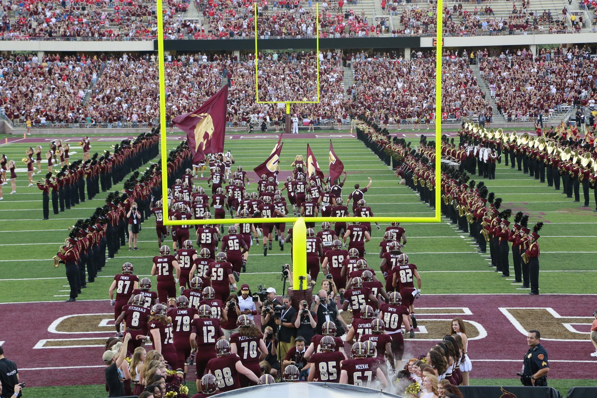 Texas State football players run onto the field inside Bobcat Stadium with the Texas State band and cheer squads tunneling them towards the goalpost as fans fill the stands.