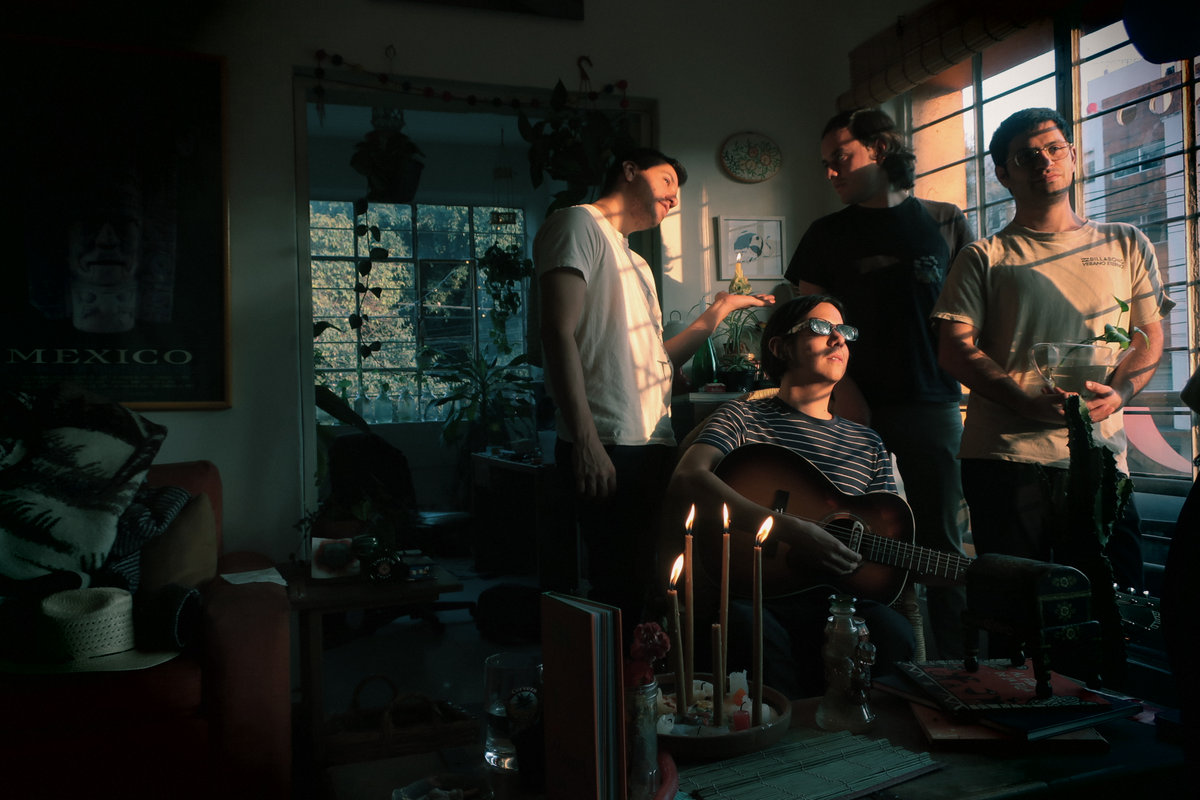 This is a group photo of the four members of Los Blenders standing in a dim room, next to a window with the sunlight shining on their faces.
