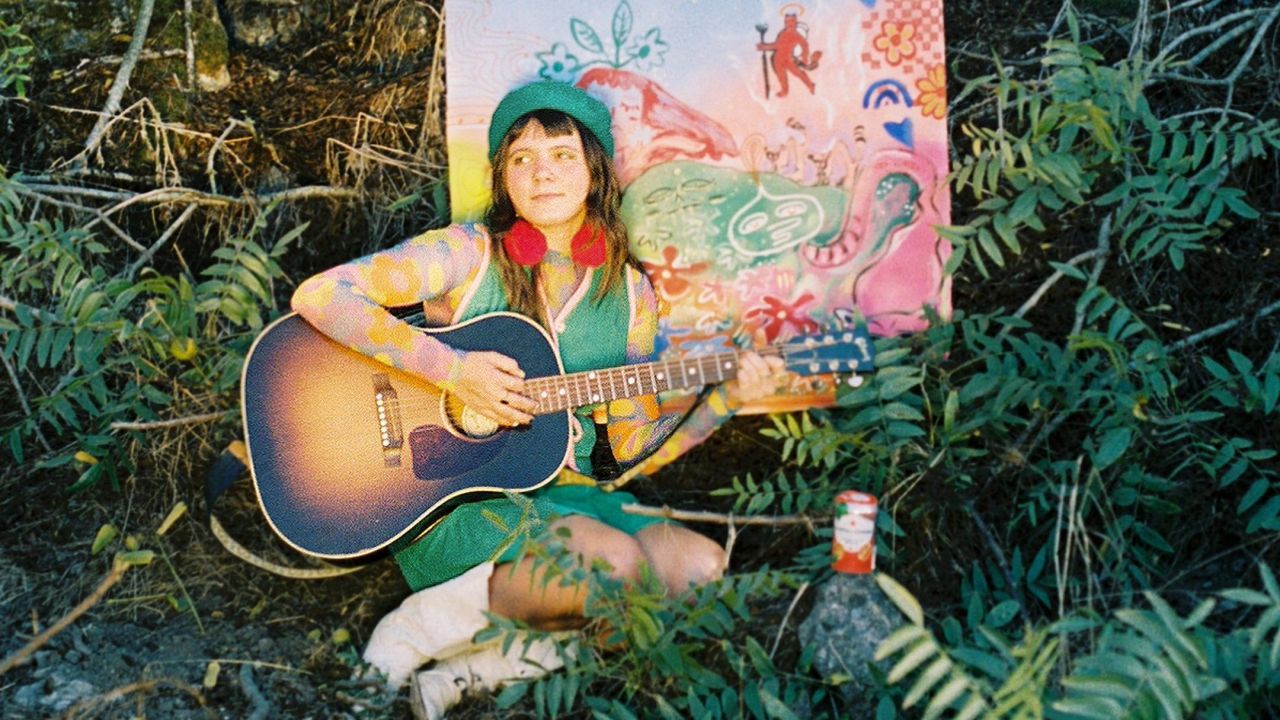 Photo of Mae Powell with acoustic guitar amidst greenery.
