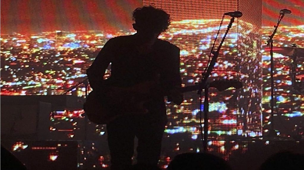The photo is a live performance of the lead singer’s shadow playing guitar and the drumist playing the drums with a projected background of city lights at night. he 1975 performing their song “Medicine” in Austin, Texas in 2017
