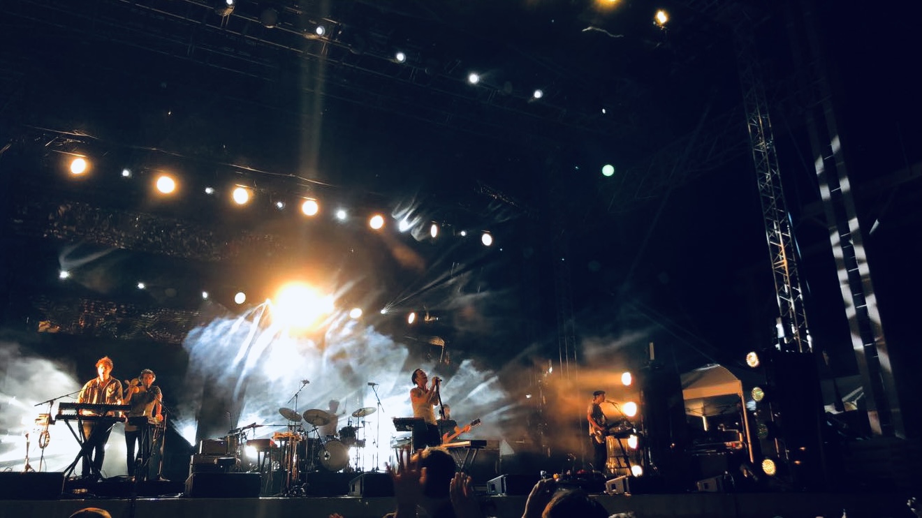 A picture of a band performing on an outdoor stage at night with stage lights shining down.