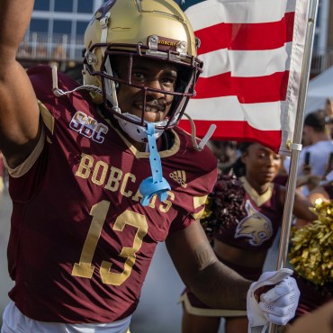 Texas State football player in Maroon jersey and gold helmet running onto the football field with an American flag with his team and cheerleaders running alongside him