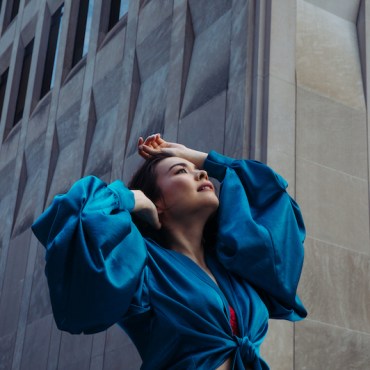 Mitski, a woman, wearing a royal blue long sleeve shirt or dress, with her hands up on her head and face, looking towards the sky with a grey building with windows in the background,