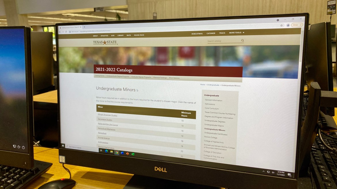 A Dell desktop monitor in the Alkek library displays a webpage from Texas State online undergraduate 2021-2022 catalog on Google Chrome. The title of the page is Undergraduate Minors and underneath is a table of offered minors listed alphabetically with a column of listed minimum hours to the right of each minor.