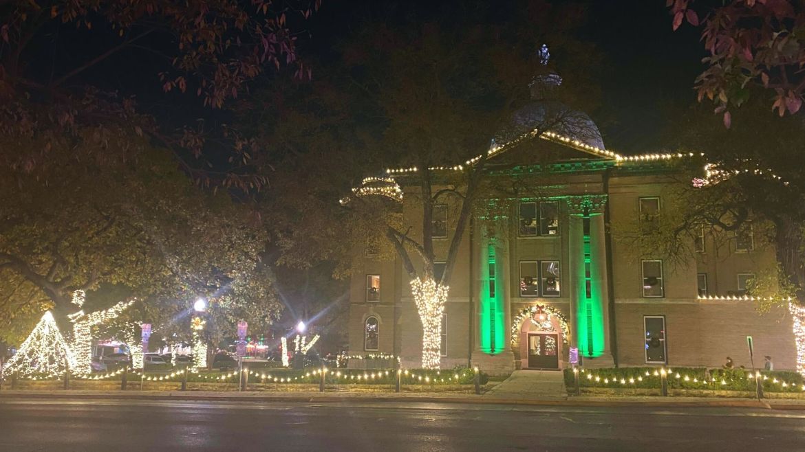 A view from East Hopkins Street, San Marcos, Texas near Kissing Alley of the Square at nighttime facing the Hays County Historic Court House. The courthouse is decorated with white string Christmas lights around the perimeter of the roof with two green LED lights shining on two pillars on each side of the north entrance also decorated with Christmas lights and a holiday wreath. Trees around the courthouse are wrapped with white string lights and the fence surrounding the property is completely lit by string lights.