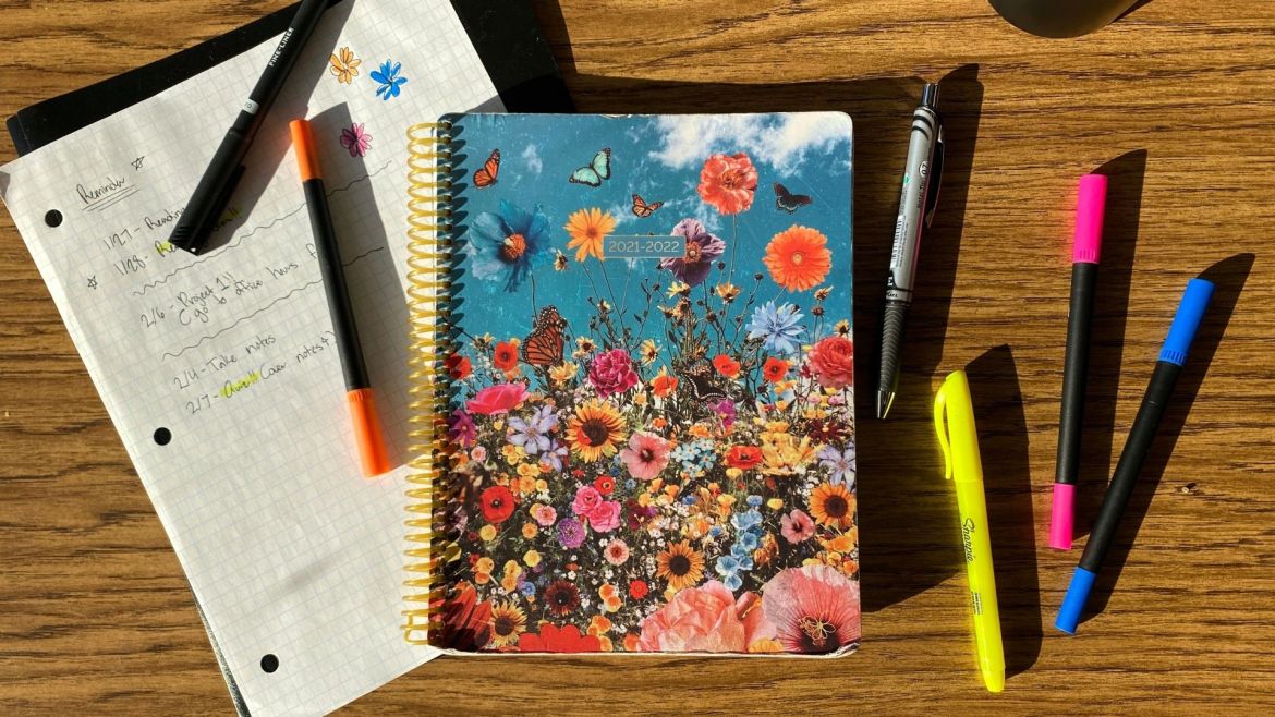 On a wooden table sits a piece of grid paper with handwritten text and small pen drawings atop a black folder and a multicolored spiral planner with wildflowers and butterflies on the cover. The planner cover reads 2021-2022. To the right of the planner is a black pen, a yellow Sharpie highlighter and pink and blue marker pens. On top of the grid paper is a black marker and an orange marker pen. In the top right corner is the edge of an opaque black water bottle.