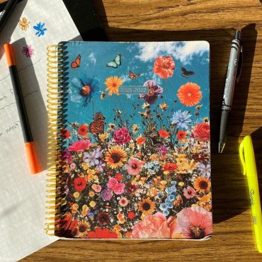 On a wooden table sits a piece of grid paper with handwritten text and small pen drawings atop a black folder and a multicolored spiral planner with wildflowers and butterflies on the cover. The planner cover reads 2021-2022. To the right of the planner is a black pen, a yellow Sharpie highlighter and pink and blue marker pens. On top of the grid paper is a black marker and an orange marker pen. In the top right corner is the edge of an opaque black water bottle.
