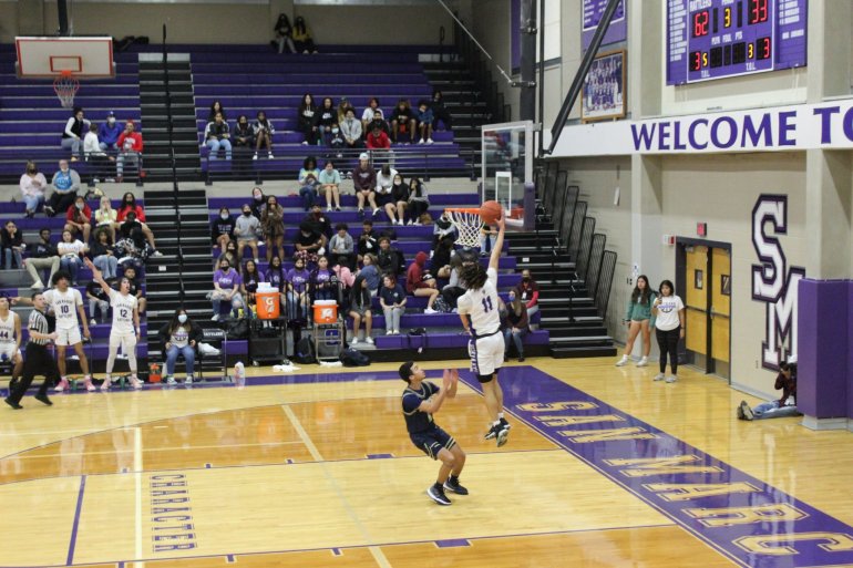 A player for the San Marcos Rattlers basketball team is shooting for a shot at the basketball goal while a player for the Lake Travis Cavaliers stands below him on the court. The court is beige with a purple stripe that reads "San Marcos". In the background, the crowd cheers from the purple bleachers.