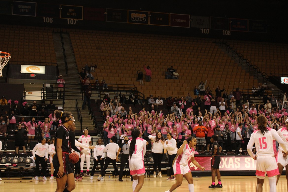 Many players in white and pink for the cancer game, with the diamond sweethearts in the back. There are three players in black on the court.
