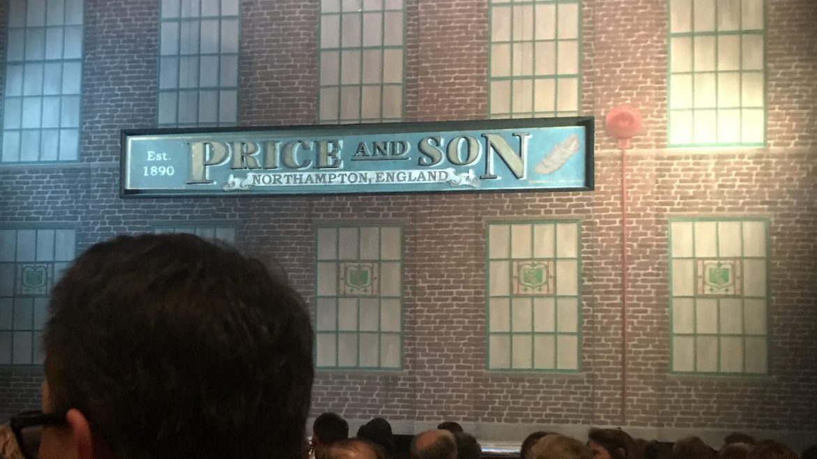 The opening set of Kinky Boots, the musical, is showcased. The set is the outside of the building of Price and Son shoe factory. There is a big green sign that says “Price and Son.” The building is brick, two stories, with ten long windows, five on each floor. The floor audience is seen in their seats.