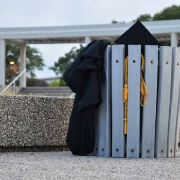A black cap and gown hang out of a gray trashcan in the park.