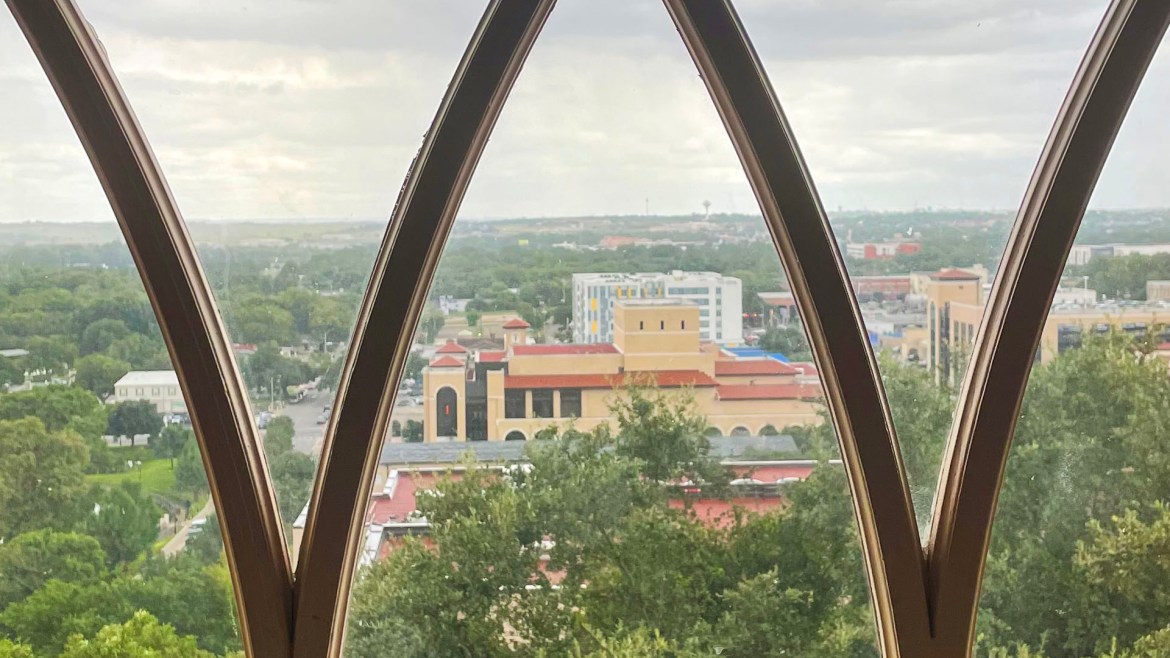 Photo is taken from a high window. In the window, there are curvy wooden lines. What is outside the window is a bird’s eye view of Texas State University. There are a lot of trees and buildings with red rooftops. In the middle of the view is a large university building with cream-colored bricks and a red rooftop. Behind that building is a tall apartment building that is white with yellow squares on the left side of it and windows spaced out throughout the building. The sky in the picture is very cloudy and grey but there is some sunlight peeking out of the clouds.