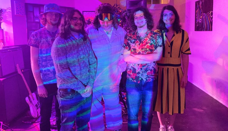 A photo of, from left to right, Kevin Schultz, Aiden Potter, Stephen Douglas, Dallas Anderson, and Leonora Tomlinson of Window Shop. They are standing in front of their musical equipment, with purple lighting. 