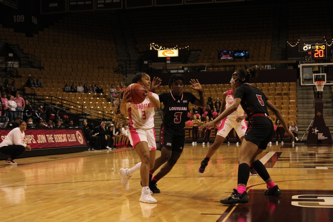 Texas State Woman's basketball player Kennedy Taylor looking to pass the ball in a pink & White uniform, with two Louisiana player in black guarding Kennedy Taylor.