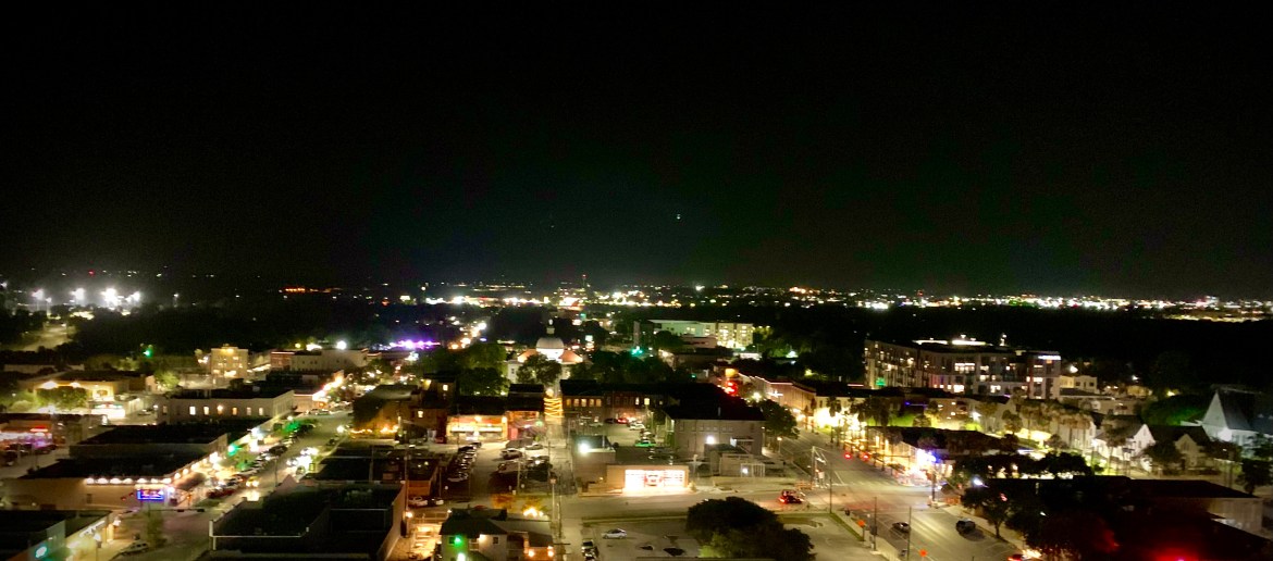An elevated view of downtown San Marcos at night. Straight ahead the lights of kissing alley and parts of the square surrounding it are visible. The skyline behind the square is also visible.