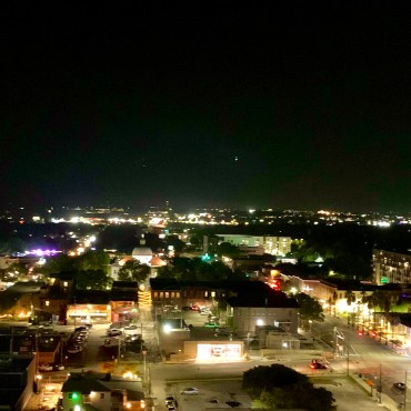 An elevated view of downtown San Marcos at night. Straight ahead the lights of kissing alley and parts of the square surrounding it are visible. The skyline behind the square is also visible.