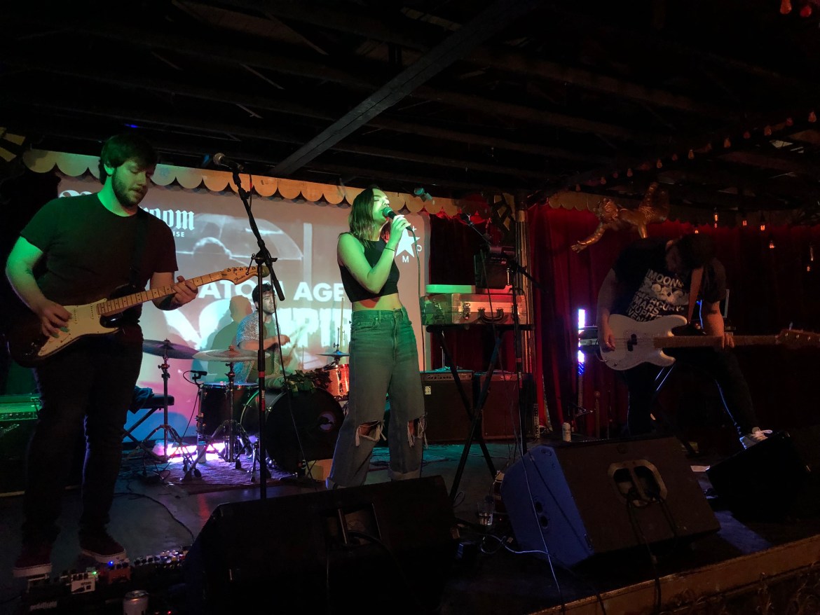 Photo features the band Chancla fight club performing on stage lit up with blue and green lights. There is a projection of some cartoons behind them. Front and center is lead singer Ashley Dawson, a bass player to her left, a guitar player to her right, and a drummer behind her.