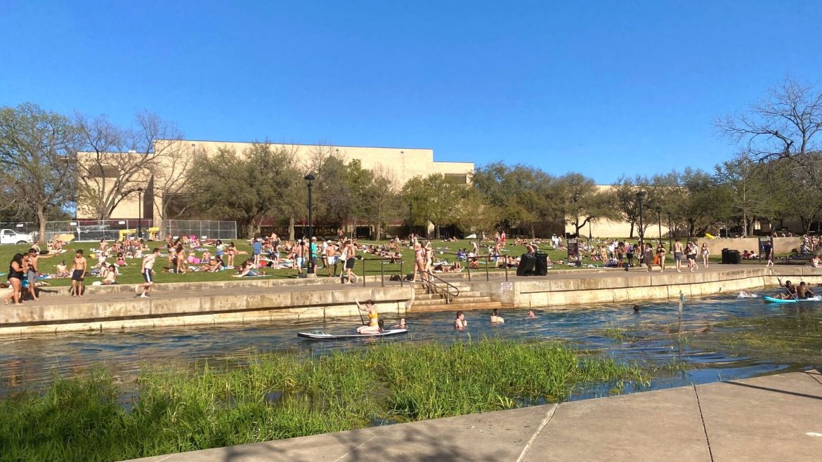 A wide view of one side of Sewell Park on a busy weekend. In the San Marcos River, there are adults on paddle boards, in canoes and swimming. There are dozens of people walking alongside the concrete and sitting on beach towels in swimsuits.