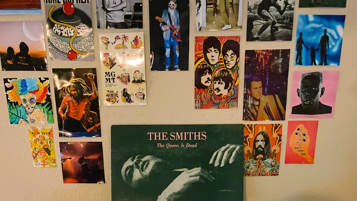 A light wooden bookshelf with crystals and a photograph on the lower shelf. A brown and black record player with a clear cover lid and two large black speakers on opposite sides is sitting on the top of the bookshelf. The vinyl cover for the album The Queen is Dead is sitting on the top of the record player facing forward. The vinyl cover is a muted dark with a person laying down and their head towards the left with pink text at the top of the album reading “The Smiths. The Queen Is Dead.” The back wall is covered in 5x7 images of various bands, artists, and album covers.
