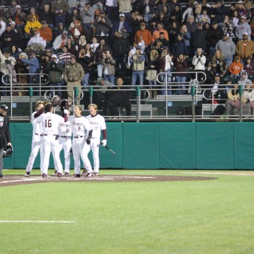 Five Texas State baseball players in a white uniform with maroon Texas State on their chest and numbers on their back. Two players tilting their helmets with a crowd in the back standing and cheering in the stands.