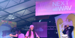 Monaleo, seen wearing her signature bright pink shirt, pink glasses and blue beanie, smiles on stage. Behind her is the two girls she brought on stage to rap with her, as well as her DJ. Behind all of them is several psychedelic tapestries and a pink flag that says Soundcloud Next Wav.