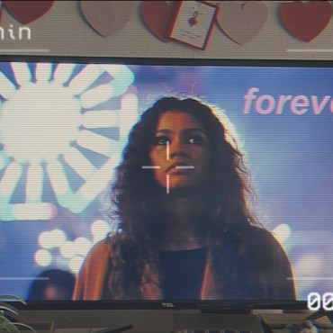 Tv screen showing Zendaya in front of a blurred bright white Ferris wheel and purple background with the word "forever" in the upper right hand corner. Above the tv are dark and light heart pink decorations with plants below the tv. On the photo is an overlay of a camera recording filter.