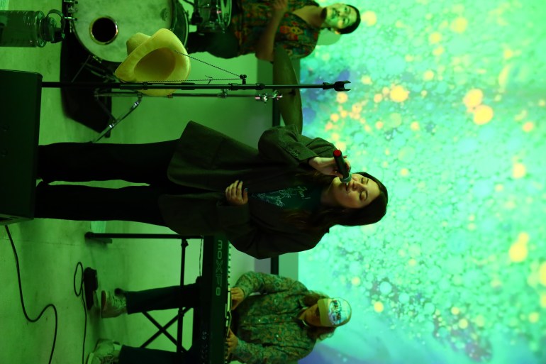 Photo features Grace Womack singing with Pianist, Luke to her left and drummer, Renee to her right. They are all lit with green lighting and there is a green, yellow, and blue tie dye image projected behind them. 