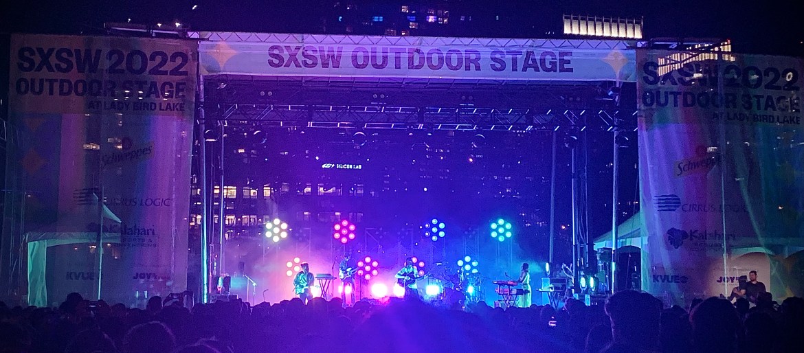 The top of the stage reads, “SXSW OUTDOOR STAGE,” with a large banner on either side that reads, “SXSW 2022 OUTDOOR STAGE AT LADY BIRD LAKE” with a list of sponsors underneath. On stage, 5-piece group Mt. Joy is performing with purple and blue lights behind them.