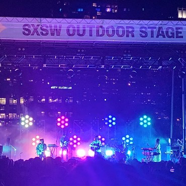 The top of the stage reads, “SXSW OUTDOOR STAGE,” with a large banner on either side that reads, “SXSW 2022 OUTDOOR STAGE AT LADY BIRD LAKE” with a list of sponsors underneath. On stage, 5-piece group Mt. Joy is performing with purple and blue lights behind them.