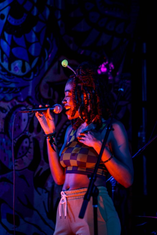 Kae from The S.O.U.L. holds a microphone and is singing facing the left of the frame. She is wearing a green checkered tank top and sweat shorts. She has a headband with 2 green puffball springs on top. She is lit by purple spotlights. 