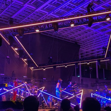 Up close image of the band Bon Iver in a large concert venue. There is a drummer, singer playing the guitar, and another performer playing the bass behind a layered keyboard. They are each surrounded by diamond diagonal lighted blue and orange light structures. Each structure consists of individual bars of light connected at points in the air and on the ground. Above the band is a square structure with double lined orange lights. Individual lights are facing the stage between the two light beams around the square. The bottom of the photogram has many people's heads as they watch the show.