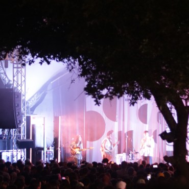 A large tree to the right is framing the stage. Three performers are seen on the stage with a purple and blue hue shining on them. A large crowd is seen before them