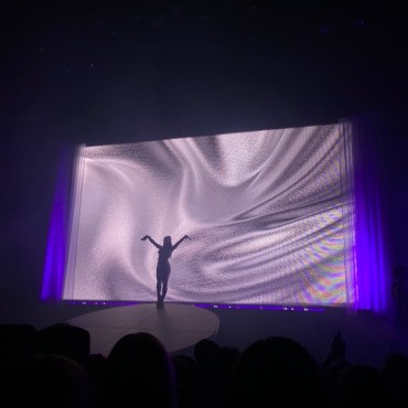 Photo features the silhouette of Pop Singer Charli XCX alone on stage, holding her arms in the air as she takes in the applause. She is standing in front of a digital projection of what seems to be silver satin.