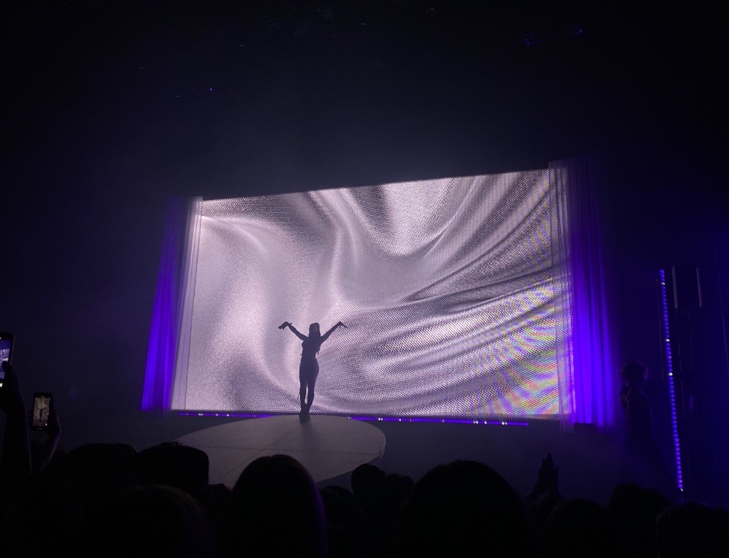 Photo features the silhouette of Pop Singer Charli XCX alone on stage, holding her arms in the air as she takes in the applause. She is standing in front of a digital projection of what seems to be silver satin.