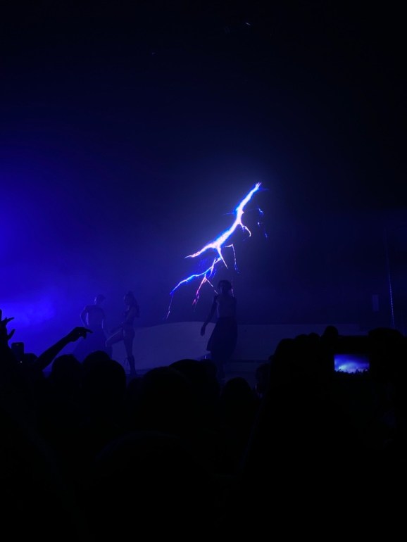 Photo features almost a pitch black stage with the tint of a blue light illuminating Charli XCX and her supporting dancers. A bolt of lightning is seen shooting down on the digital screen behind them.