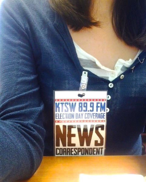 The image is a close up on a blue shirt with a tag pinned to it that says “KTSW 89.9 FM, Election Day Coverage” at the top. At the bottom, it says “News Correspondent” in bold black letters. 