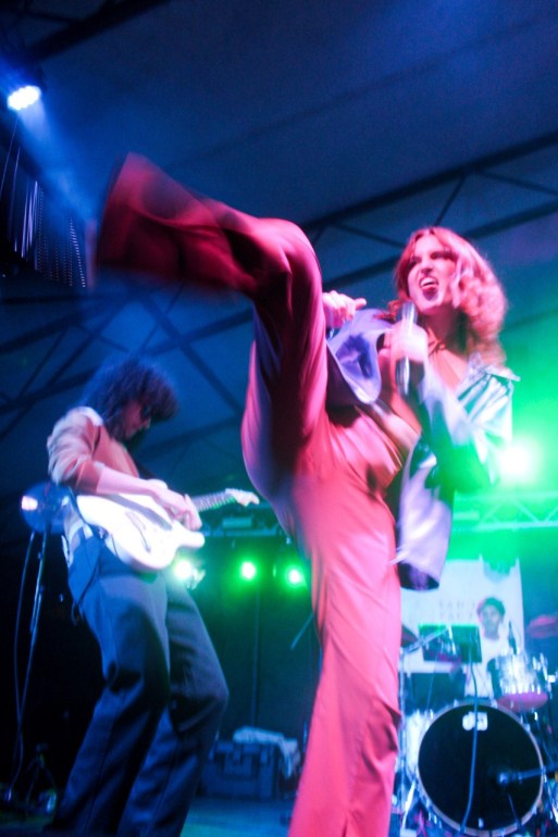 Katie Gavin, in an orange jumpsuit topped with a purple blazer, does a high kick onstage with her tongue out. Naomi McPherson and drummer background the photo. 