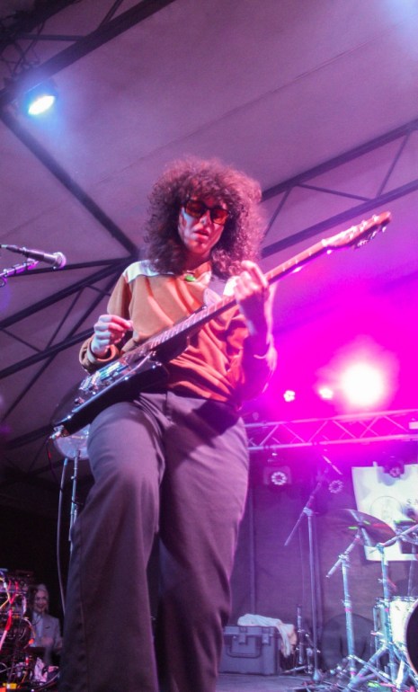 Illuminated by red stage lighting, Naomi’s orange-tinted sunglasses complement their orange long-sleeved top paired with brown pants. Naomi plays an electric guitar balanced on their right leg.