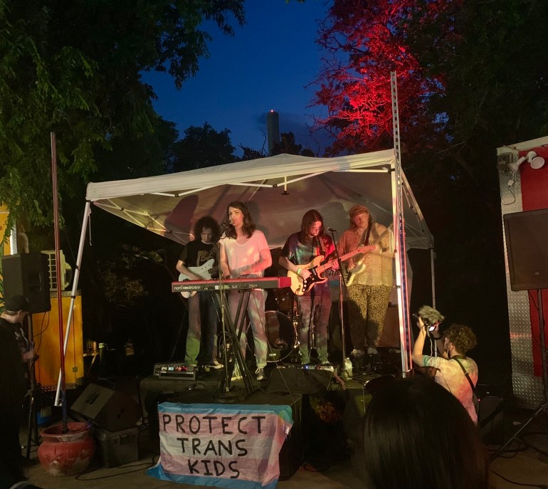 The photo features a small stage with a white tent overhead, and a pink, blue, and white flag on the front that reads, “Protect Trans Kids.” There are trees behind the stage that are backlit with red and pink, and a dark blue sky behind the trees.