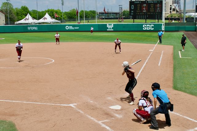 A hitter is at the plate ready for the ball to be thrown. The setting is a softball field.