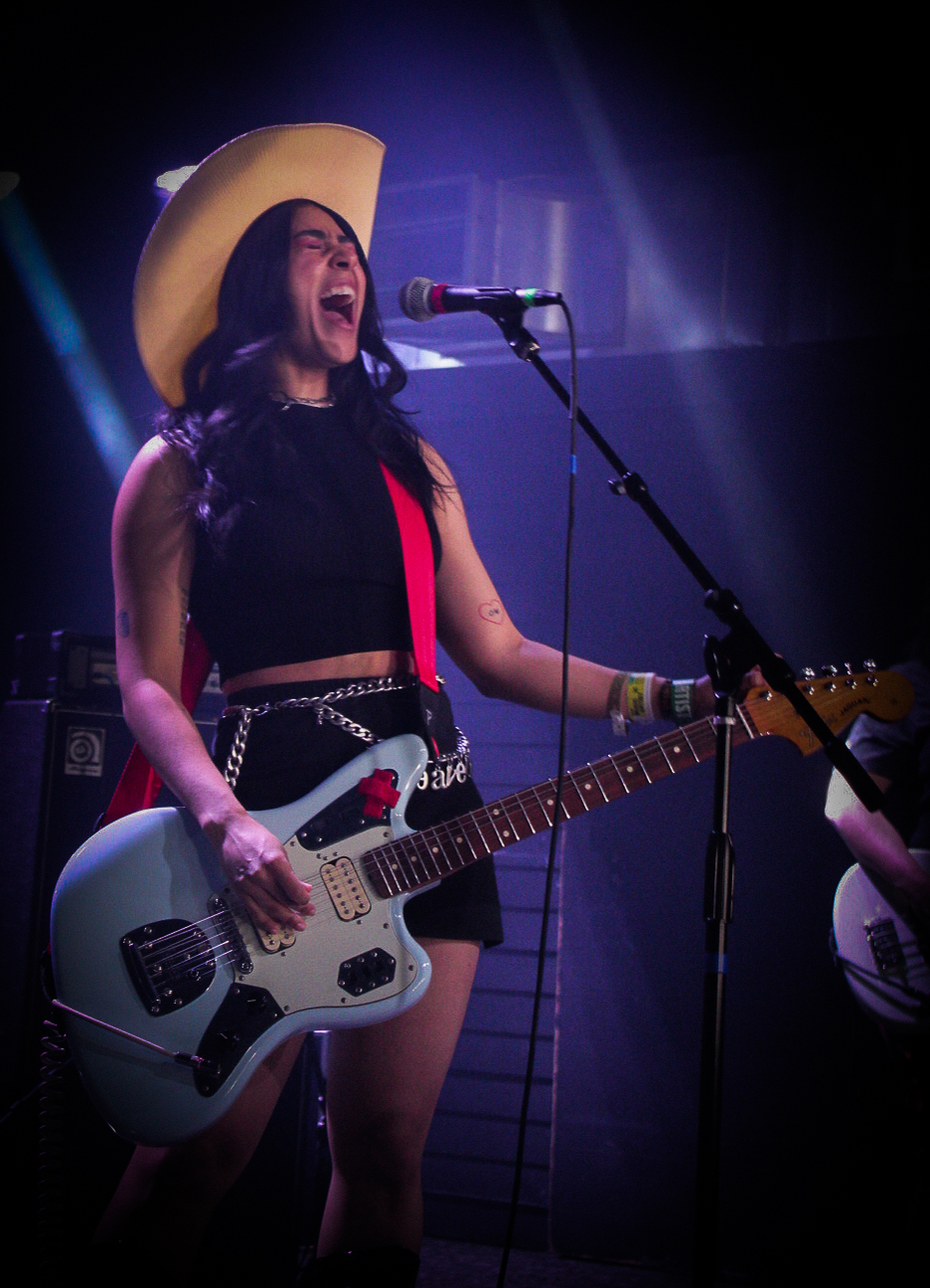  Mia Berrin wears a black two-piece with a silver chain belt, and a white cowboy hat, while playing a powder blue electric guitar.