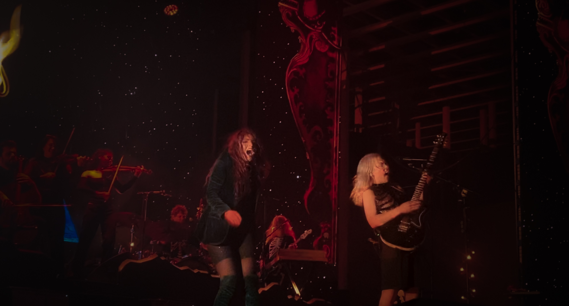 Phoebe Bridgers with a black electric guitar and Haley Dahl in royal blue blazer, backgrounded by red stage lighting.