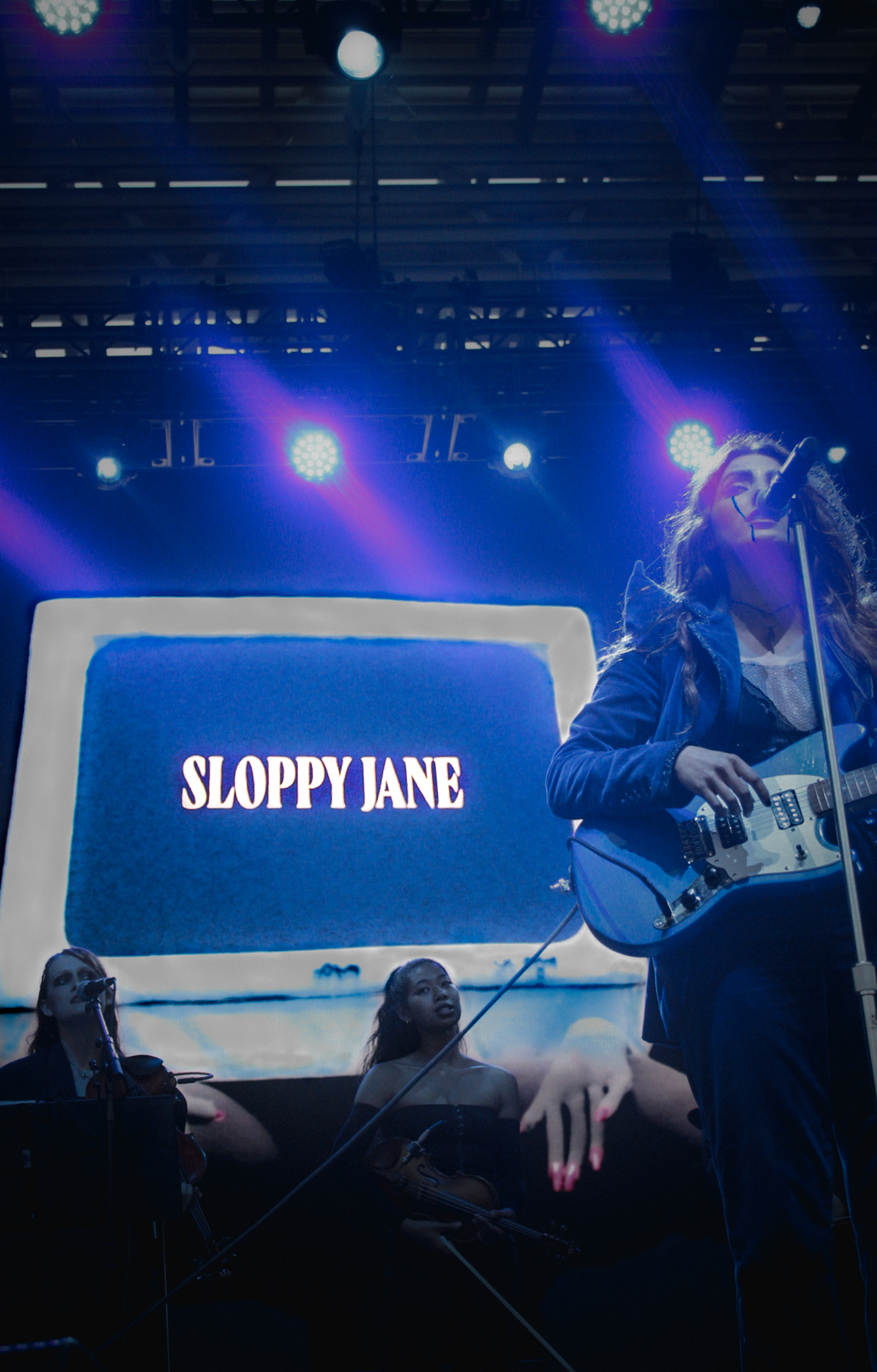 Haley Dahl with a blue electric guitar, illuminated by a blue glow and backgrounded by a white television, reading “SLOPPY JANE.”