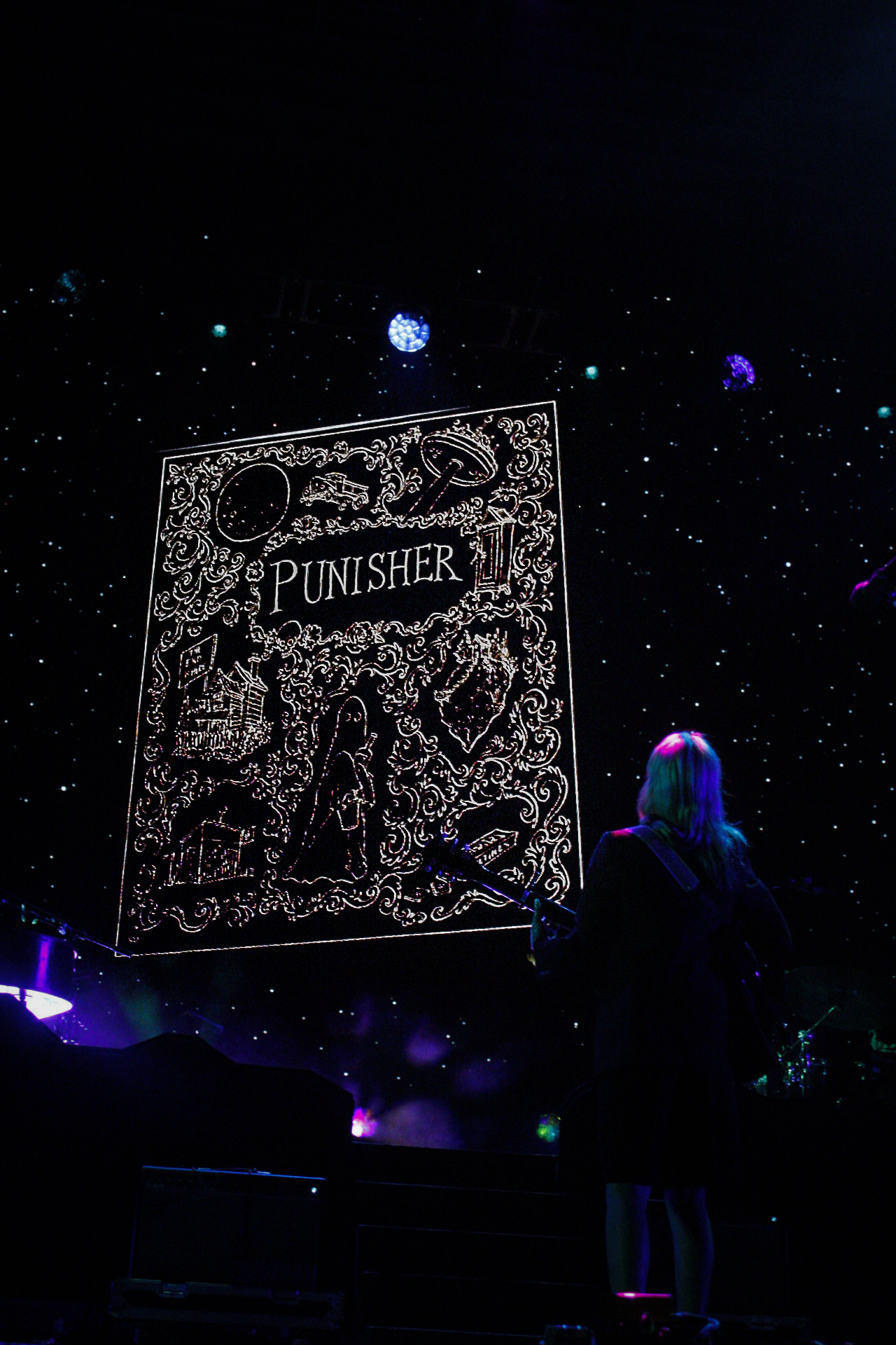 A graphic of a Punisher storybook appears with white sketches of lyrical elements – including a moon, ghost, “the end is near” sign. Phoebe Bridgers faces the screen holding a guitar.