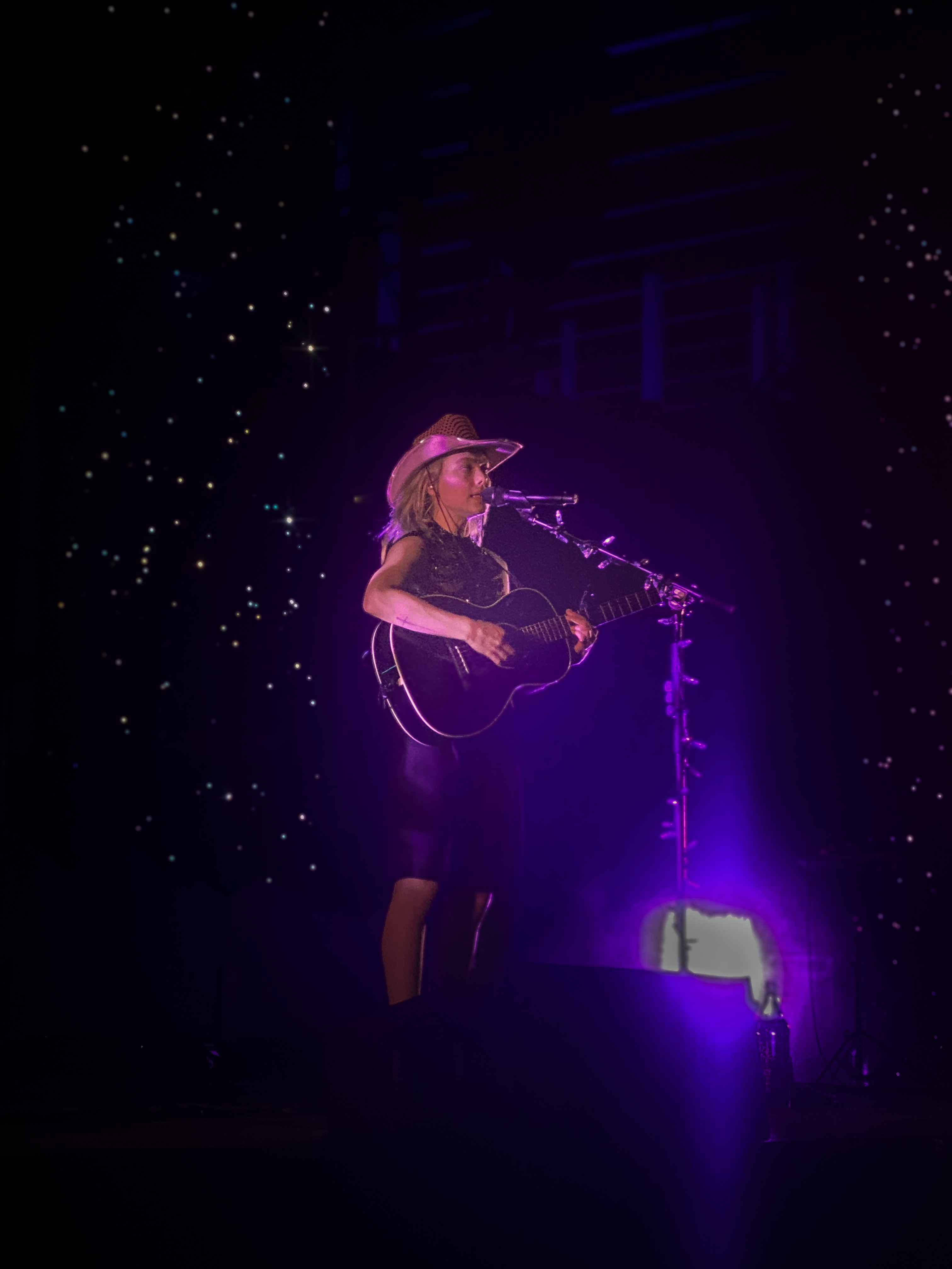 Phoebe Bridgers in a pink cowboy hat with a black acoustic guitar, backgrounded by a starry sky. 