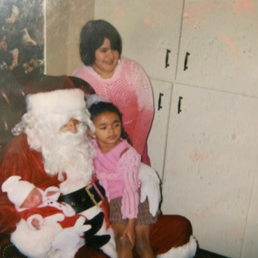 Three young children, all at different ages, take a holiday picture with Santa Clause. Standing to the far right, one girl with short dark hair is dressed in pink and white. Next to her, another young girl is seen dressed in pink and brown clothing. A baby dressed in a red and white Santa Clause outfit sits next to the girl in the middle, being held by the man in the Santa Clause outfit.